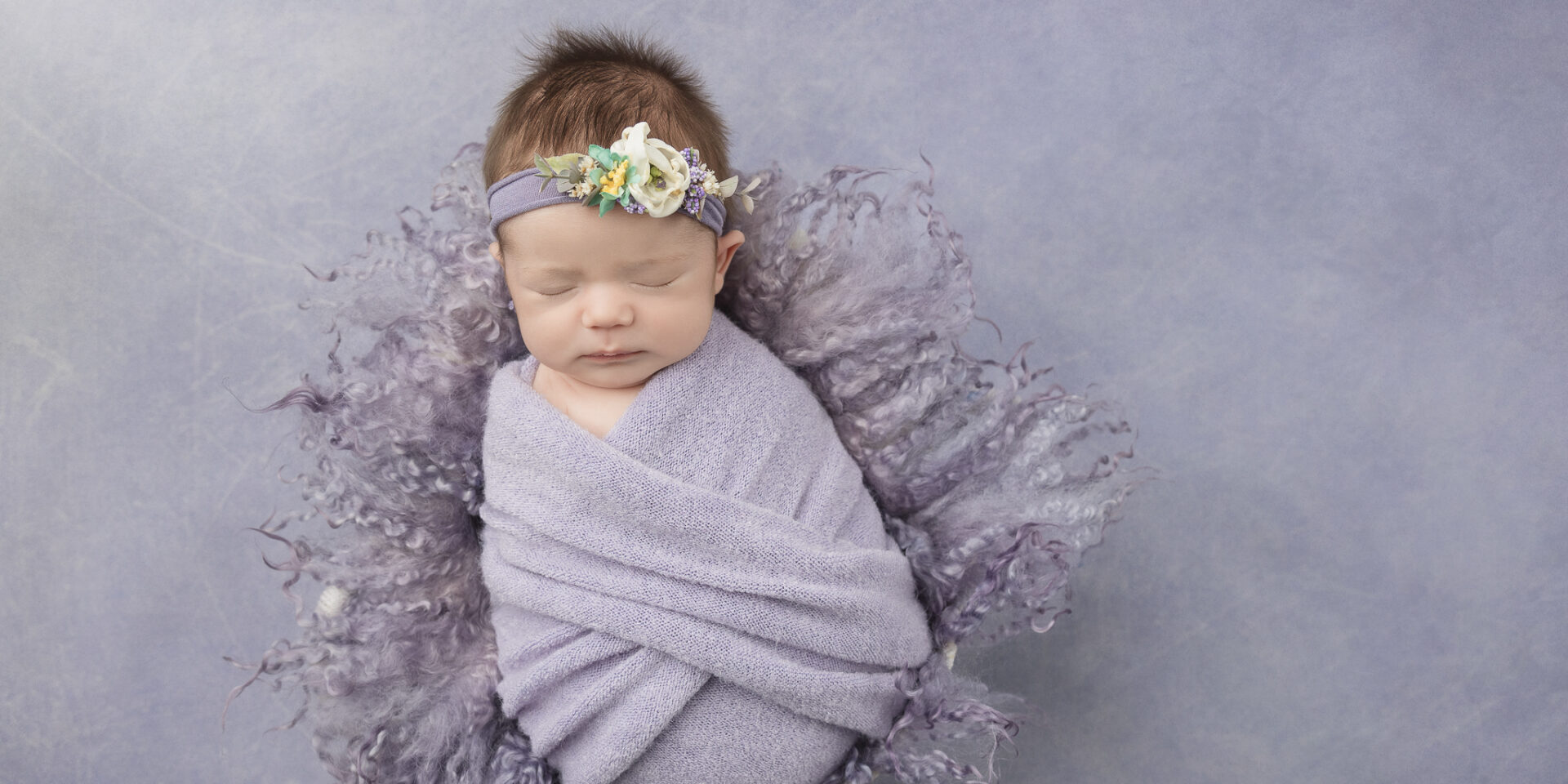 Newborn baby in purple swaddle with floral headband on purple background