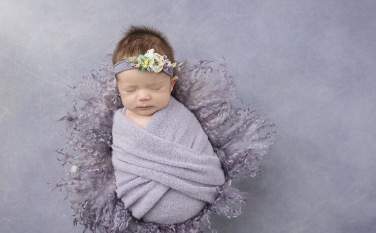 Newborn baby in purple swaddle with floral headband on purple background