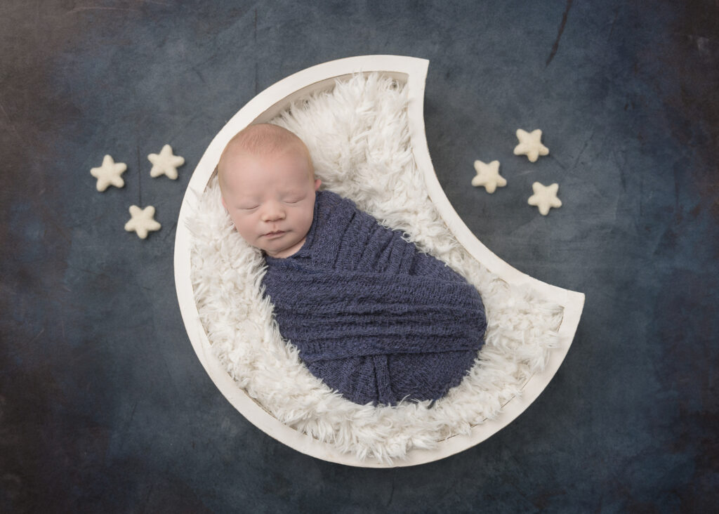 Newborn baby in blue wrap in white moon-shaped basket on blue background with white stars