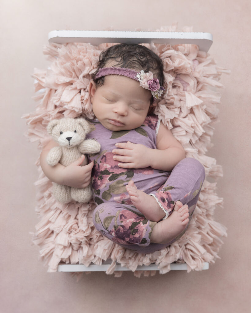 Sleeping newborn baby in tiny bed on pink fluff holding a teddy bear