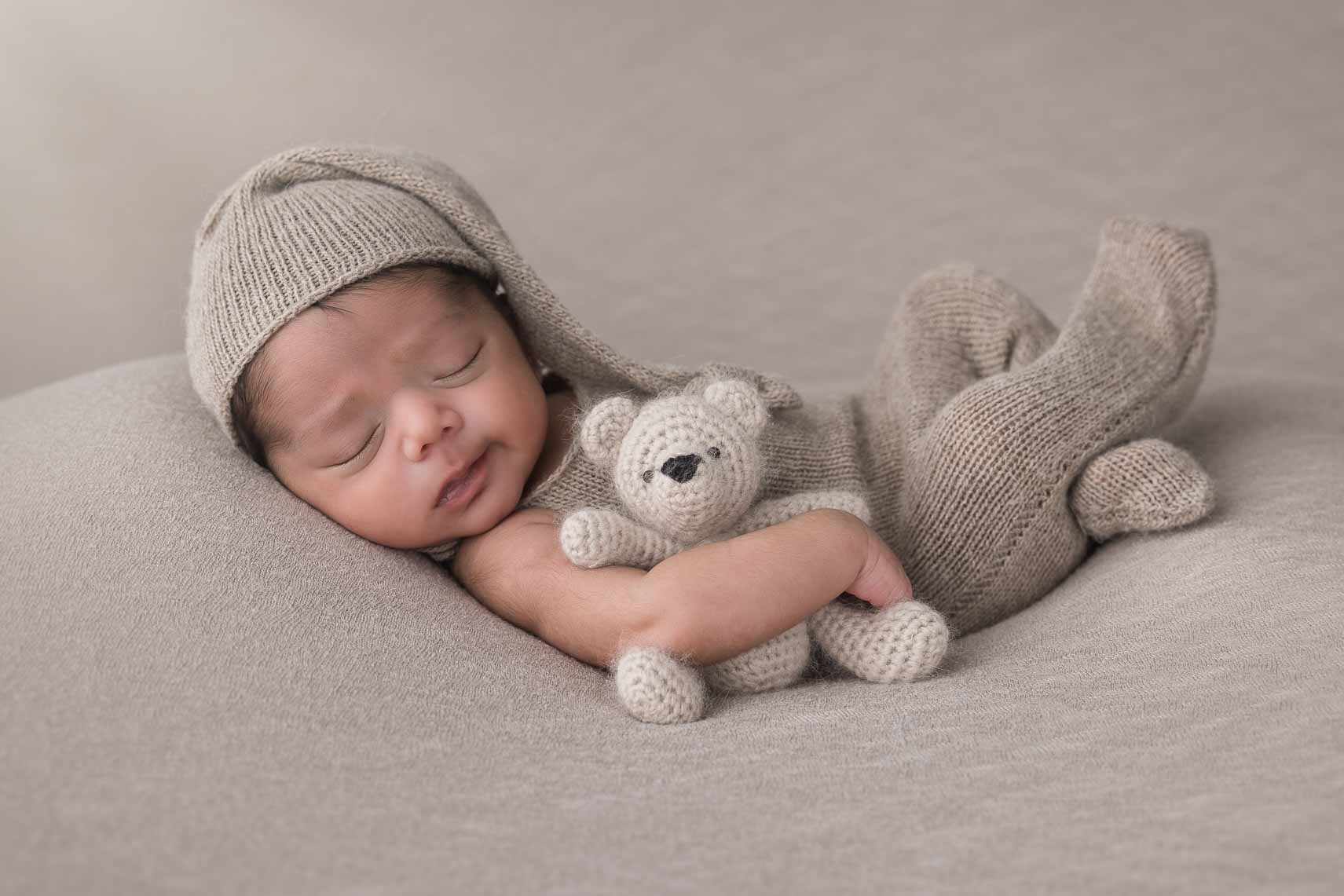 A newborn baby boy in a brown outfit with a hat on is laying on his back holding a teddy bear.