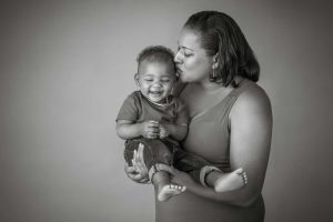 A black and white picture of a mother holding her toddler whiles she kisses the side of his forehead.