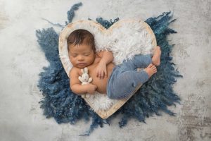 A picture from a newborn session where a little boy in blue pants is lying in a heart shaped wood bowl while holding a little bunny.