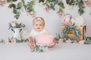 A one year old girl is sitting behind her pink cake posed in her floral themed birthday photo session setup.
