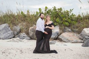 A well dress couple is posed in front of the rocks and sea oats at Jetty Park in Florida during their maternity photo session.