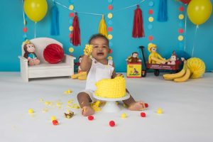 A little boy is holding a fist full of birthday cake during his Curious George themed cake smash session.