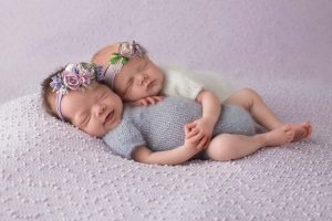 Twin girls are posed lying down sleeping together. The twin on the bottom has a big smile on her face.