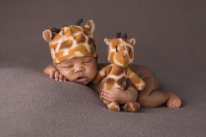 A little boy is posed lying on a blanket wearing a giraffe hat while he's holding a giraffe stuffed animal.