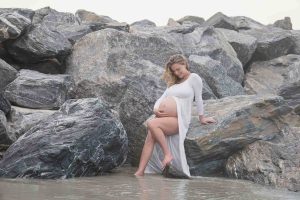 A pregnant woman in a white dress with her belly showing is posed sitting on the large rock wall at Jetty Park on the Space Coast in Florida.