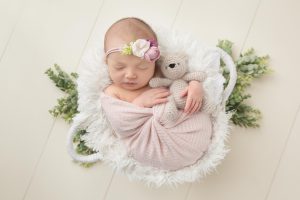 A baby girl is posed holding a teddy bear while laying in a basket prop.