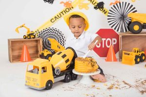A little boy in a construction themed photo session setup is using a large spoon to dig into his cake.