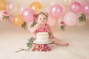 A little girl with cake on her face is sitting behind a cake with a balloon garland behind her during a birthday portrait session.