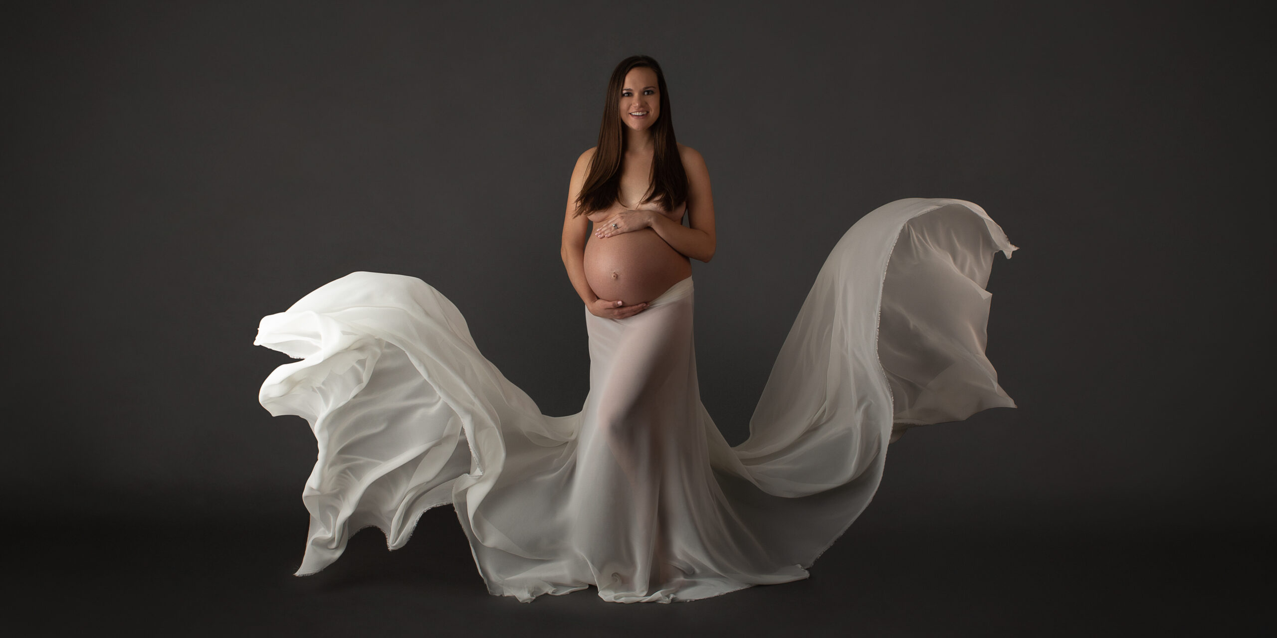 A topless pregnant woman is posing with a white skirt made of flowing fabric floating by her sides.