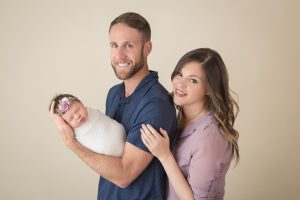A tall man is holding is newborn daughter in his arms while his wife is standing behind him hugging onto him.