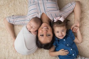 A mother is posed lying on the floor with her toddler girl & her newborn son.