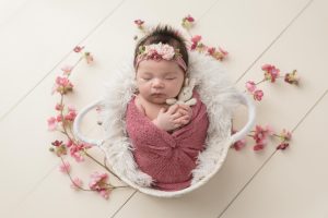 A sleeping newborn with dark hair is posed in a basket surrounded by pink flowers while she holds a little bunny.