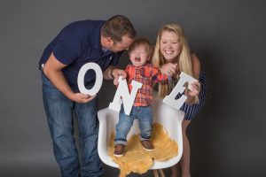 A crying toddler is standing on a chair while the parents are laughing trying to get a photo of them holding the letters o-n-e during a cake smash photoshoot.