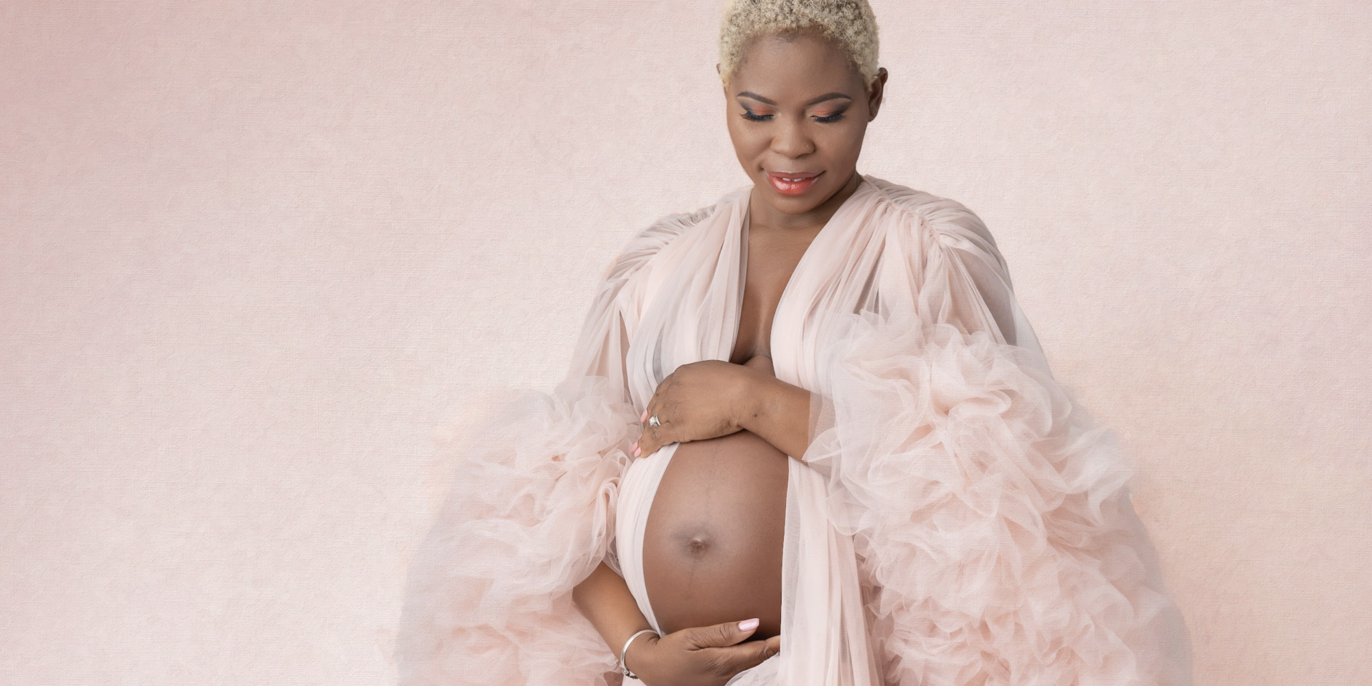 Pregnant woman on pink backdrop in pink robe, holding belly and looking down