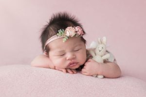 A newborn girl with super fluffy dark hair is laying on a pink blanket holding a bunny.