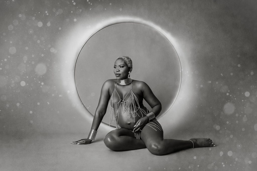Contemporary black & white picture of a woman dressed only in a decorative chain necklace, sitting on the floor with circle effects in the background.