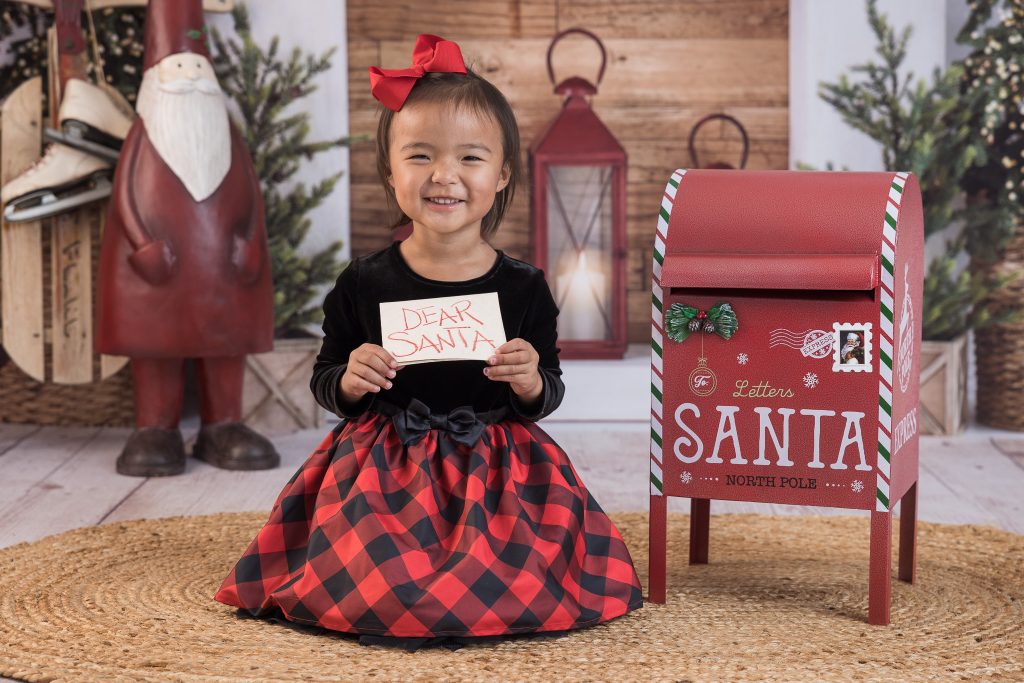Adorable Asian girl sitting on the floor posing for a Christmas picture. She's holding a Dear Santa sign and is sitting next to a Letters to Santa mailbox.