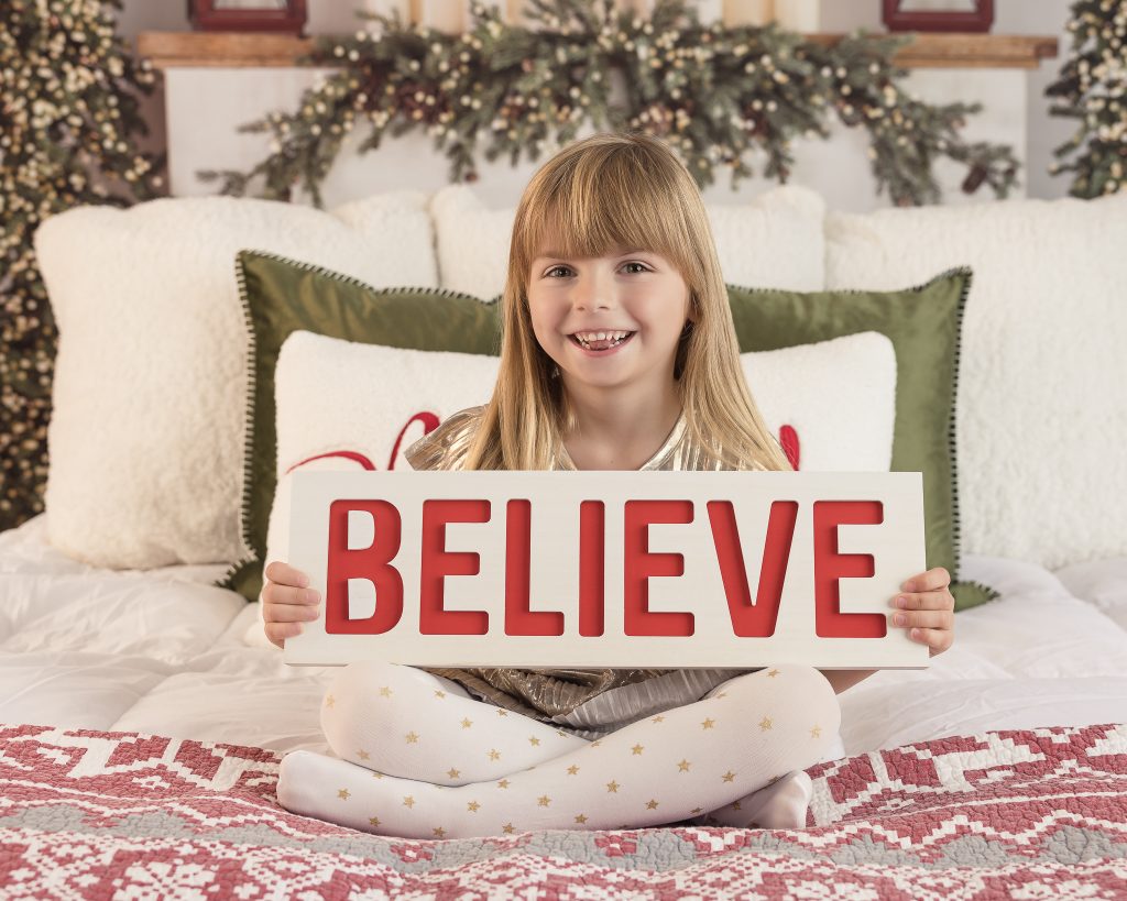 Little girl sitting on a bed holding a believe sign posing for a Christmas photo.