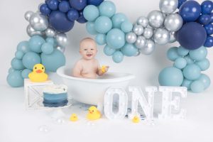 A photo of a baby during their cake smash session sitting in a bath tub surrounded by balloons, ducks and a one sign.