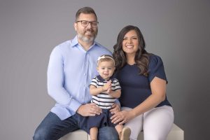 A couple and their 1 year old daughter sitting for a portrait coordinated in blue outfits.