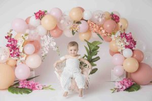 One year old girl sitting in a photography studio in a chair with an arch of balloons and flowers behind her.