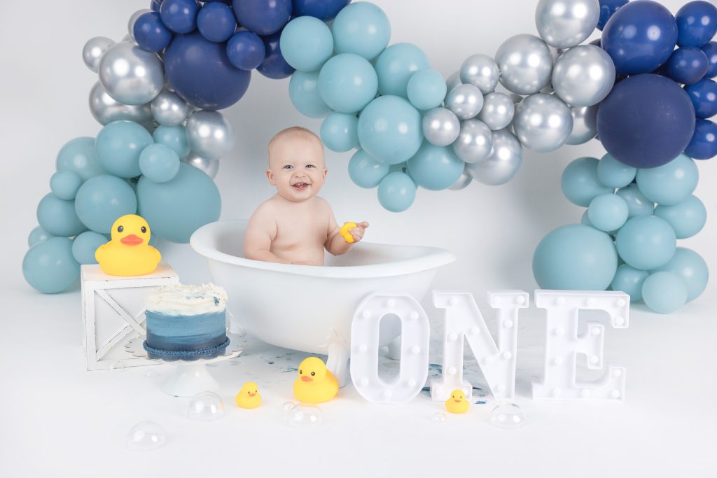 A baby sitting in a white bathtub surrounded by blue and silver balloons. The baby is smiling and holding a yellow rubber duck. There is a small blue cake, several rubber ducks, and a decorative sign with the word 'ONE' in front of the tub.