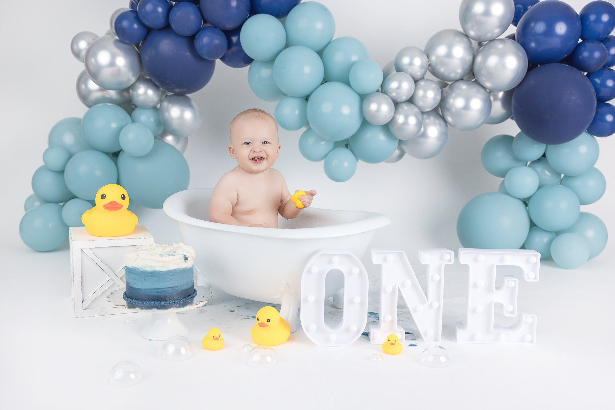 A baby sitting in a white bathtub surrounded by blue and silver balloons. The baby is smiling and holding a yellow rubber duck. There is a small blue cake, several rubber ducks, and a decorative sign with the word 'ONE' in front of the tub.